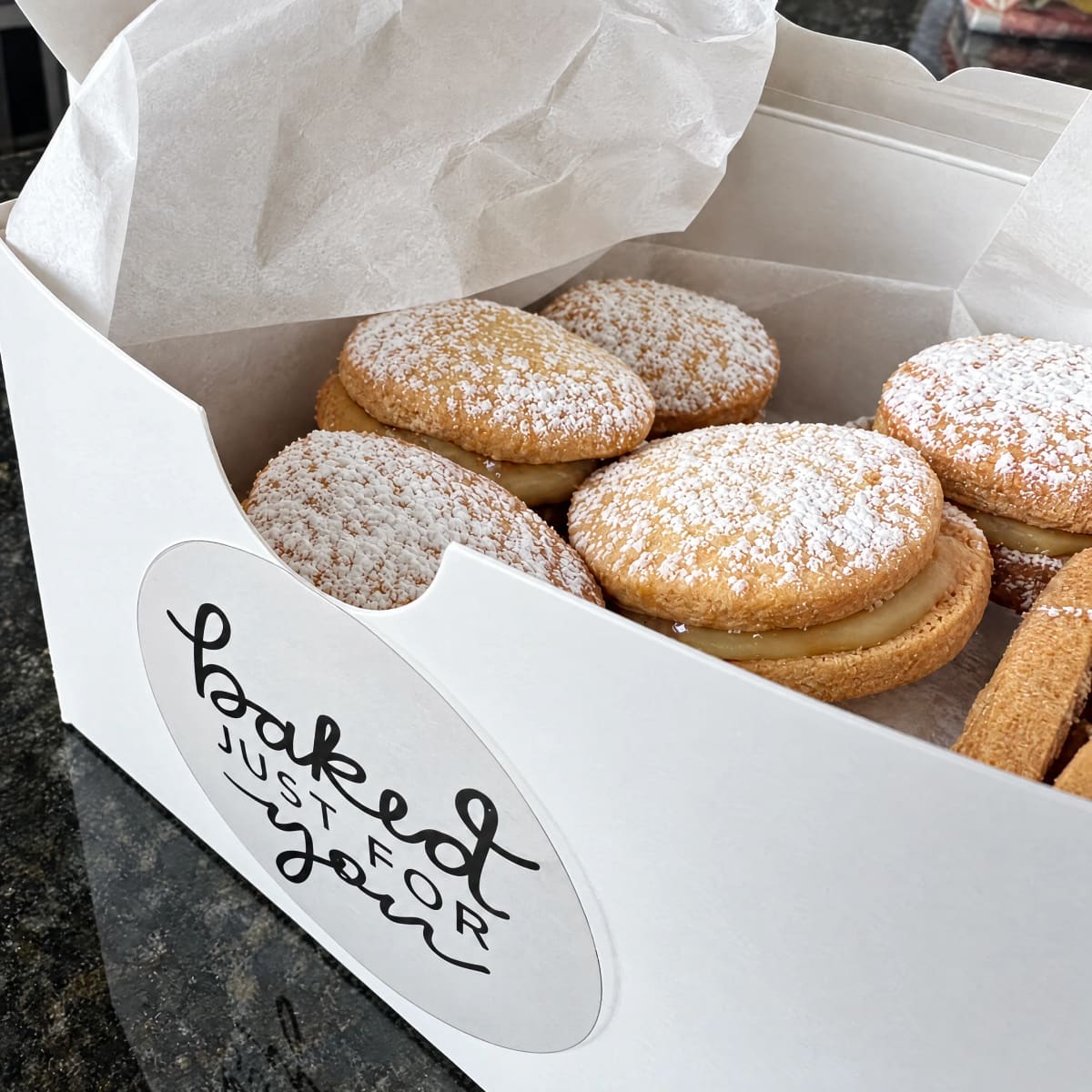 An open gift box of powdered-sugar alfajores labeled 'Baked Just For You'
