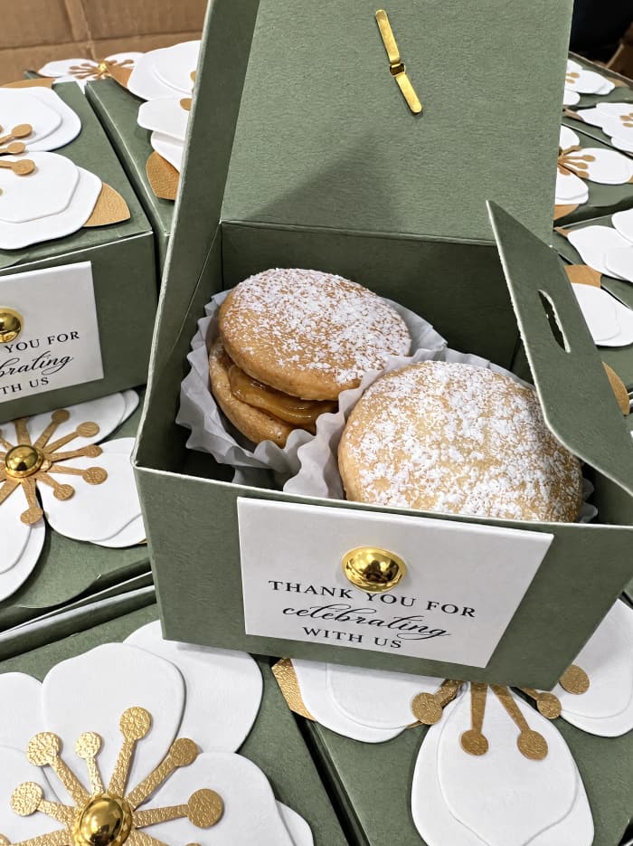 An elegant sage-green favor box with a 'Thank You For Celebrating With Us' card and alfajores inside